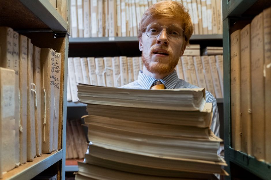 Man with glasses holding a stack of documents in a library archive, surrounded by shelves of files.